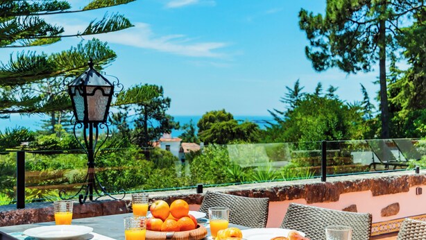 An outdoor dining area with a table set for breakfast, including croissants and orange juice, under a large orange umbrella. - Estoril Royal Atlantic Villa (Cascais)