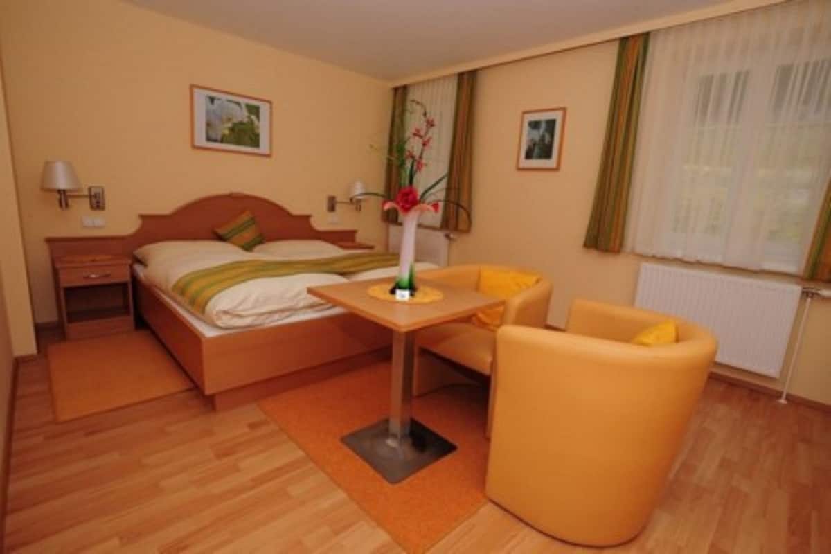 Una habitación de hotel con cama, mesita de noche, lámpara, silla y un jarrón con flores.
