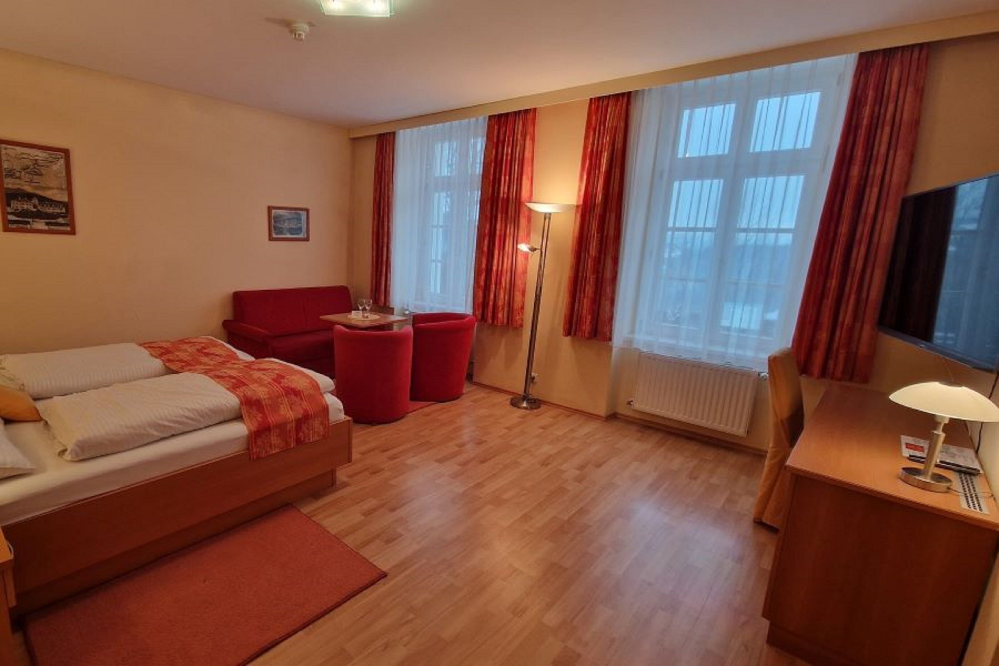A hotel room with a bed, a red armchair, a small table with a lamp, and a TV on a wooden cabinet.