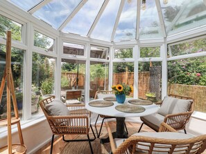 A conservatory with a round table and wicker chairs, surrounded by large windows. - The Stables Barn (Arundel)