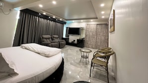A modern hotel room with a bed, two reclining chairs, a small round table, a TV, and a bathroom with a mirror and sink. - Anand Resort - A luxury Resort Nashik (Nashik)