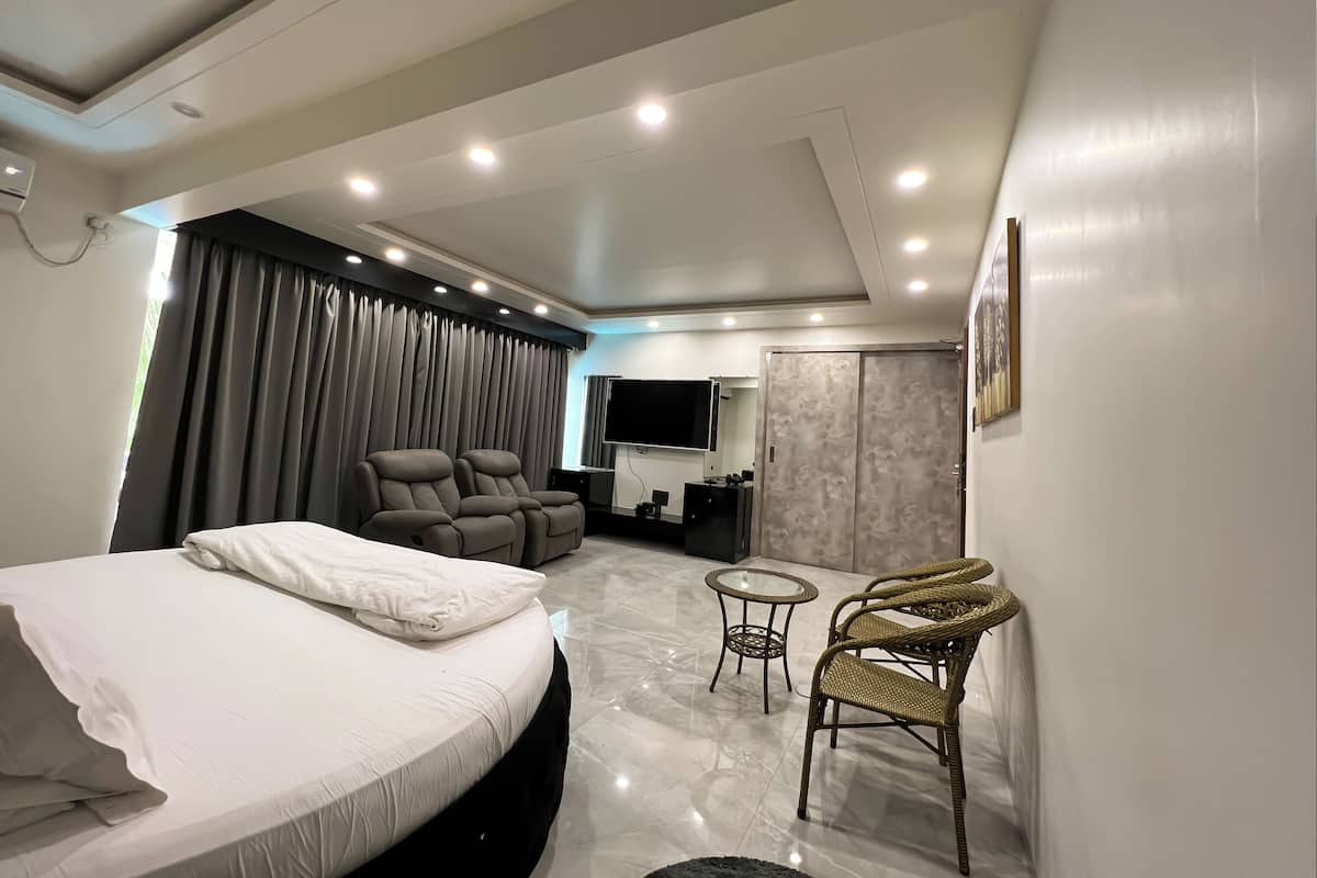 Una habitación de hotel moderna con una cama, dos sillones reclinables, una mesita redonda, un televisor y un baño con espejo y lavamanos.