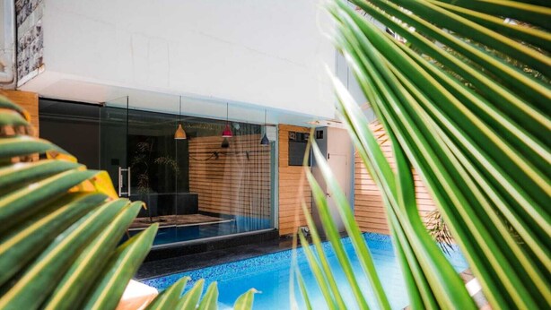 A modern outdoor pool area with a glass enclosure, wooden deck, and tropical plants. - Anand Resort - A luxury Resort Nashik (Nashik)