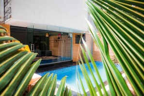 A modern outdoor pool area with a glass enclosure, wooden deck, and tropical plants.