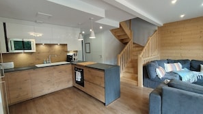 A modern kitchen with wooden cabinets, a built-in microwave, and a coffee maker. There is a wooden staircase leading to another level. A blue sofa with patterned cushions is visible in the foreground.
