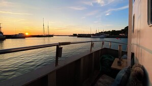 Auringonlaskunäkymä veneen kannelta, jossa on kaide, pöytä ja tuoli sekä pieni koira.