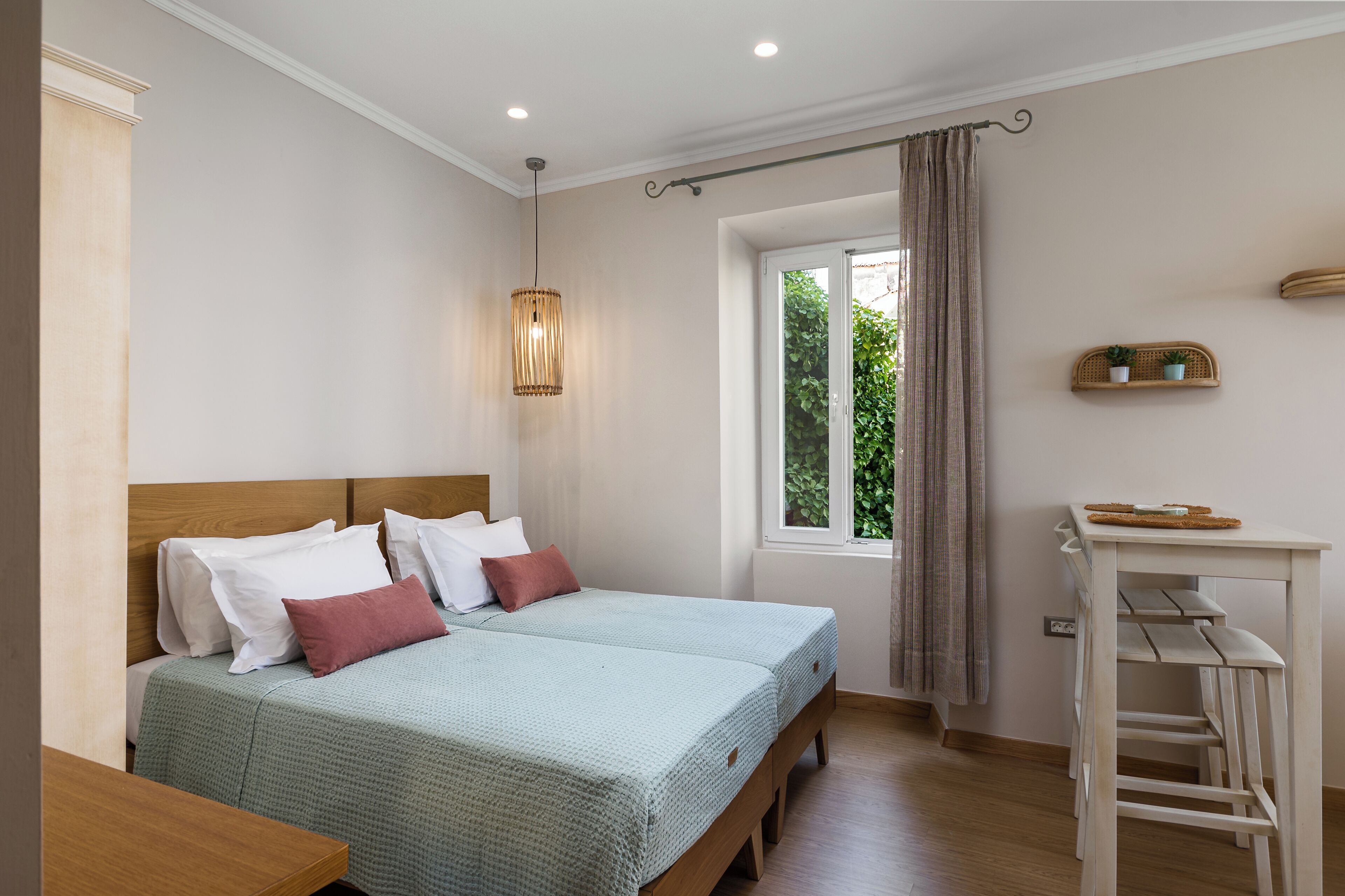 Ein Doppelbett mit Holz-Kopfstück, zwei Nachttische, eine Hängeleuchte, ein Fenster mit Vorhängen und eine kleine Ablage mit Pflanzen.