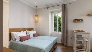 Ein Doppelbett mit Holz-Kopfstück, zwei Nachttische, eine Hängeleuchte, ein Fenster mit Vorhängen und eine kleine Ablage mit Pflanzen.