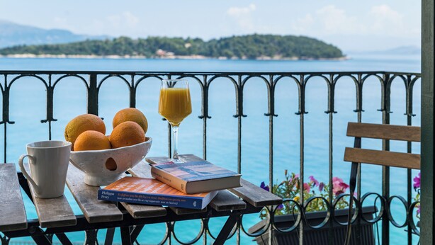 Una mesa con un cuenco de naranjas, un vaso de zumo de naranja, un libro y una taza, frente a una vista panorámica de agua y tierra.