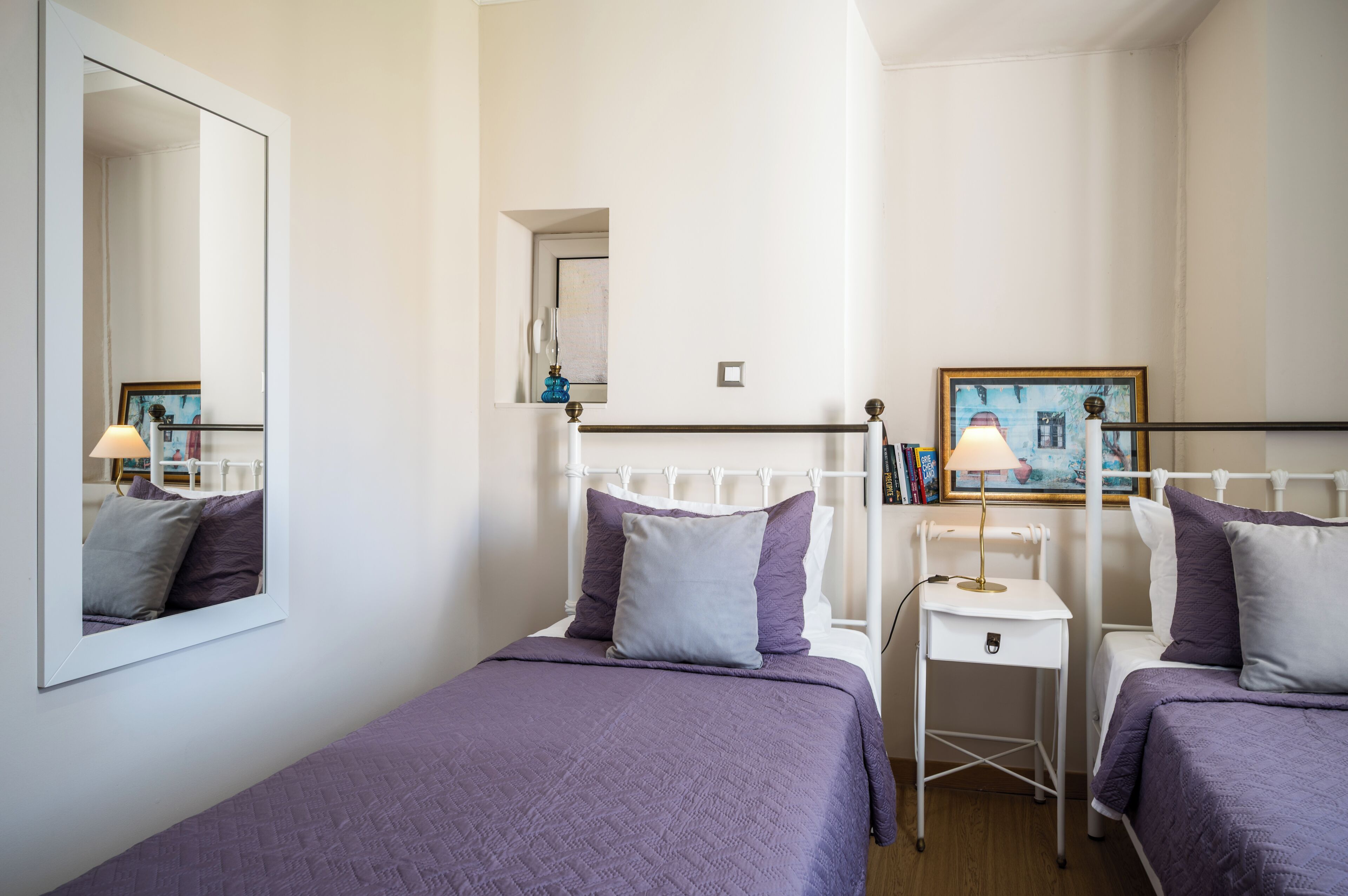 A bedroom with two beds, a bedside table with a lamp, a mirror, and a painting on the wall.