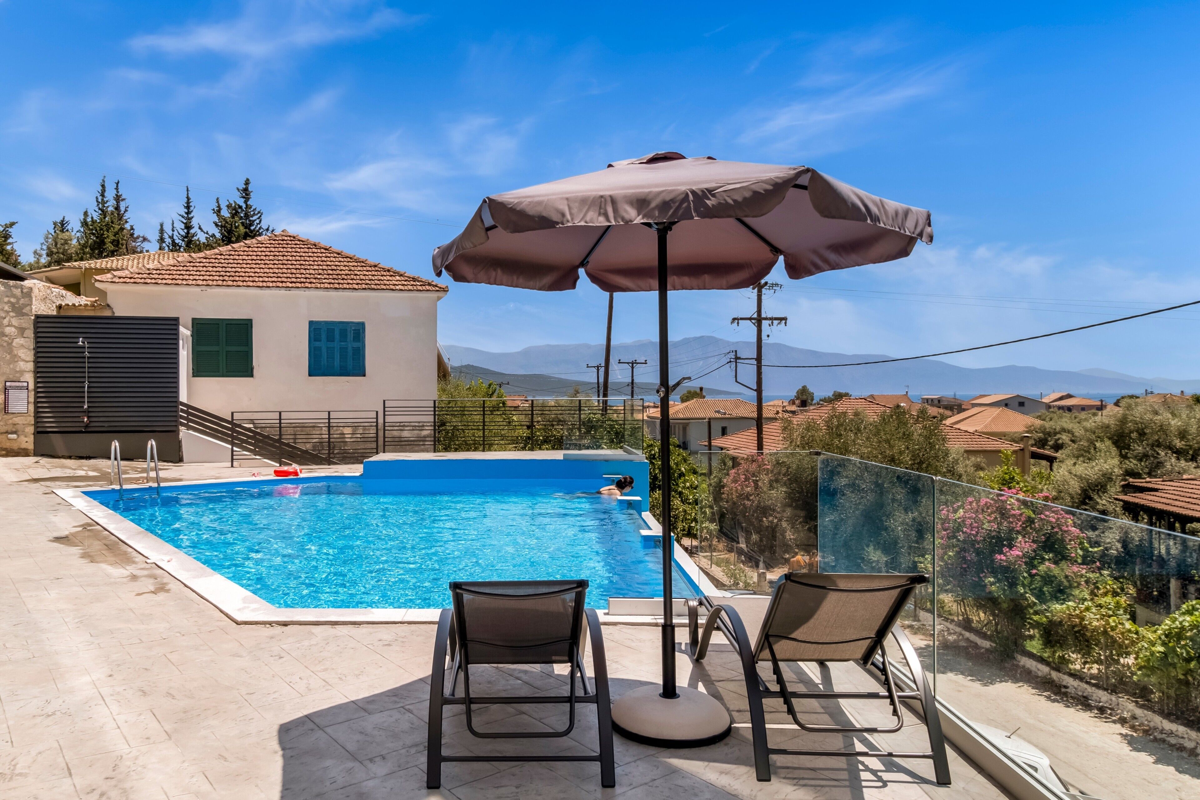 A swimming pool with two chairs and an umbrella, surrounded by a house with a tiled roof and a view of mountains.