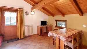 Un intérieur en bois comprenant une table à manger, un téléviseur posé sur un meuble en bois, une fenêtre avec des rideaux et un radiateur.