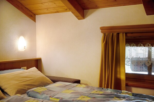 A bedroom with a bed, a nightstand, a wall-mounted light, and a window with curtains.