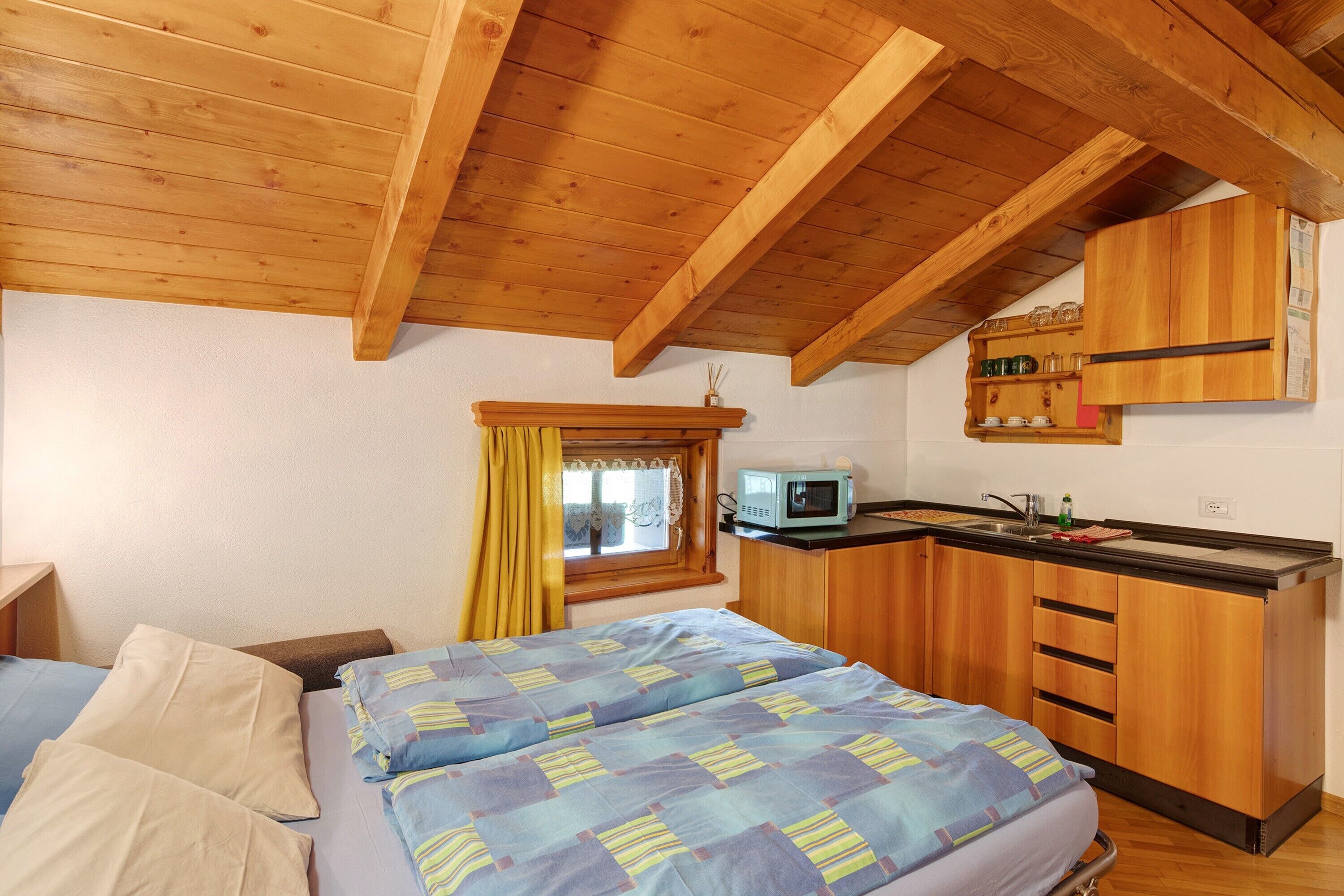 A compact room with a bed, a kitchenette, and a window with curtains.