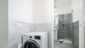 A compact laundry room with a front-loading washing machine, a glass shower enclosure, and a visible towel rack. - Holiday Home 'Casa Manu' with Private Terrace, Wi-Fi and Air Conditioning (Assemini)
