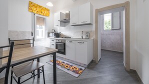 A modern kitchen with white cabinets, a built-in oven, and a coffee maker. There is a dining table with chairs and a rug with a sunflower pattern. - Holiday Home 'Casa Manu' with Private Terrace, Wi-Fi and Air Conditioning (Assemini)