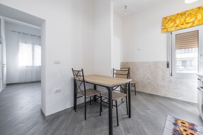 A dining area with a wooden table and four chairs, a window with blinds, and a door leading to another room. - Holiday Home 'Casa Manu' with Private Terrace, Wi-Fi and Air Conditioning (Assemini)