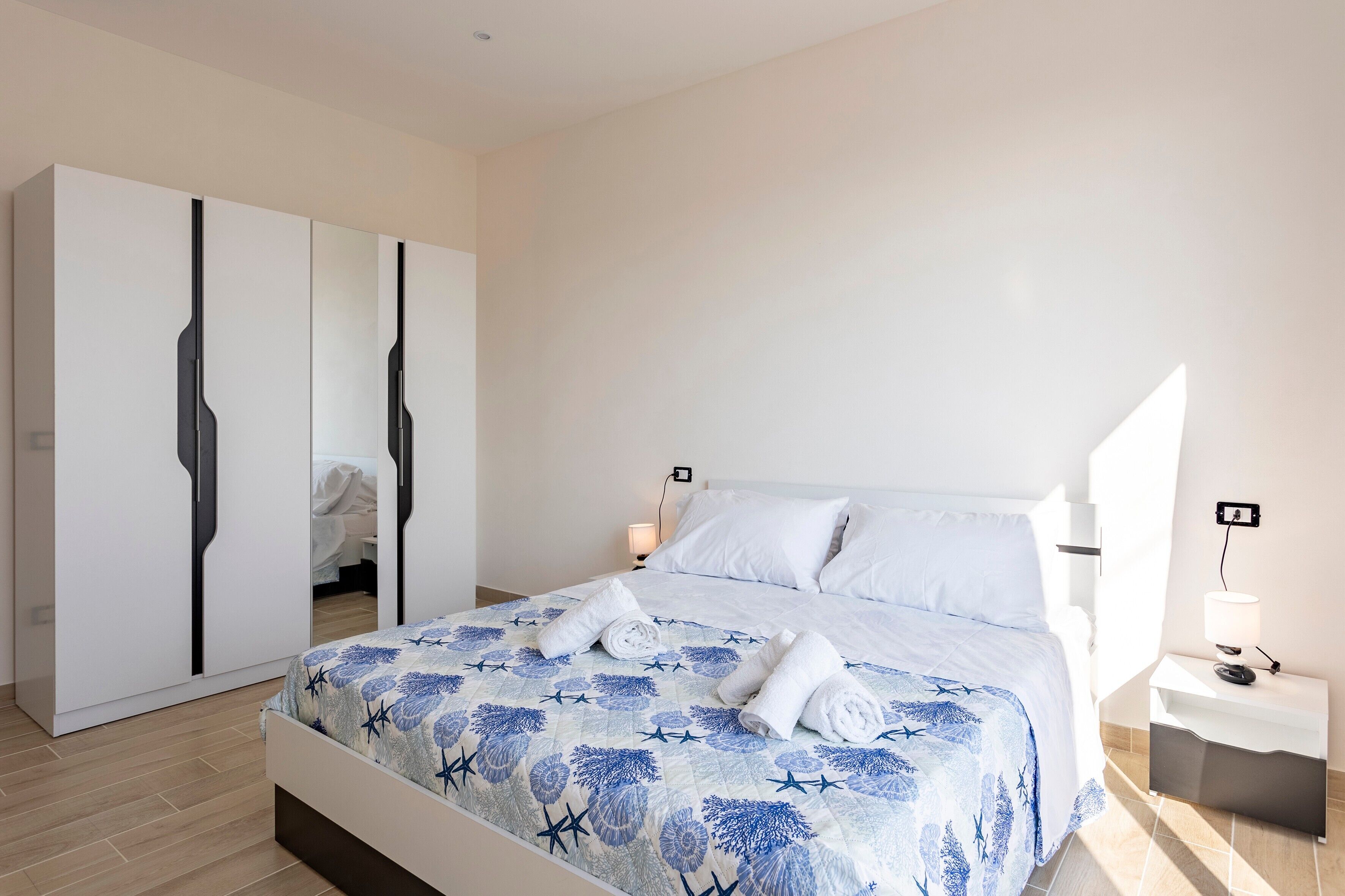 A neatly arranged bedroom with a bed, a wardrobe, a nightstand, and a wall-mounted lamp.
