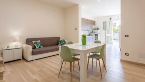 A modern living room with a sofa, a dining table with chairs, a small side table with a lamp, and a kitchen in the background. - Holiday Home "Sefy Mare a Selinunte" with Sea View, Balcony & Wi-Fi (Marinella)