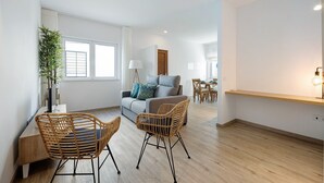 Een moderne woonkamer met een bank, twee rieten fauteuils, een eettafel en een groot raam.