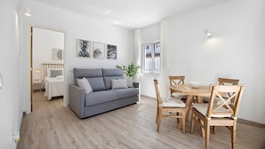 Un salon moderne avec un canapé gris, une table à manger en bois et des chaises. Un mur blanc est décoré d’œuvres d’art encadrées, et une fenêtre est agrémentée de rideaux.