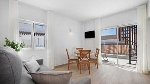Un salon moderne avec un canapé gris, une table à manger en bois et des chaises, un téléviseur à écran plat fixé au mur, et un balcon offrant une vue sur un toit recouvert de tuiles et un escalier.