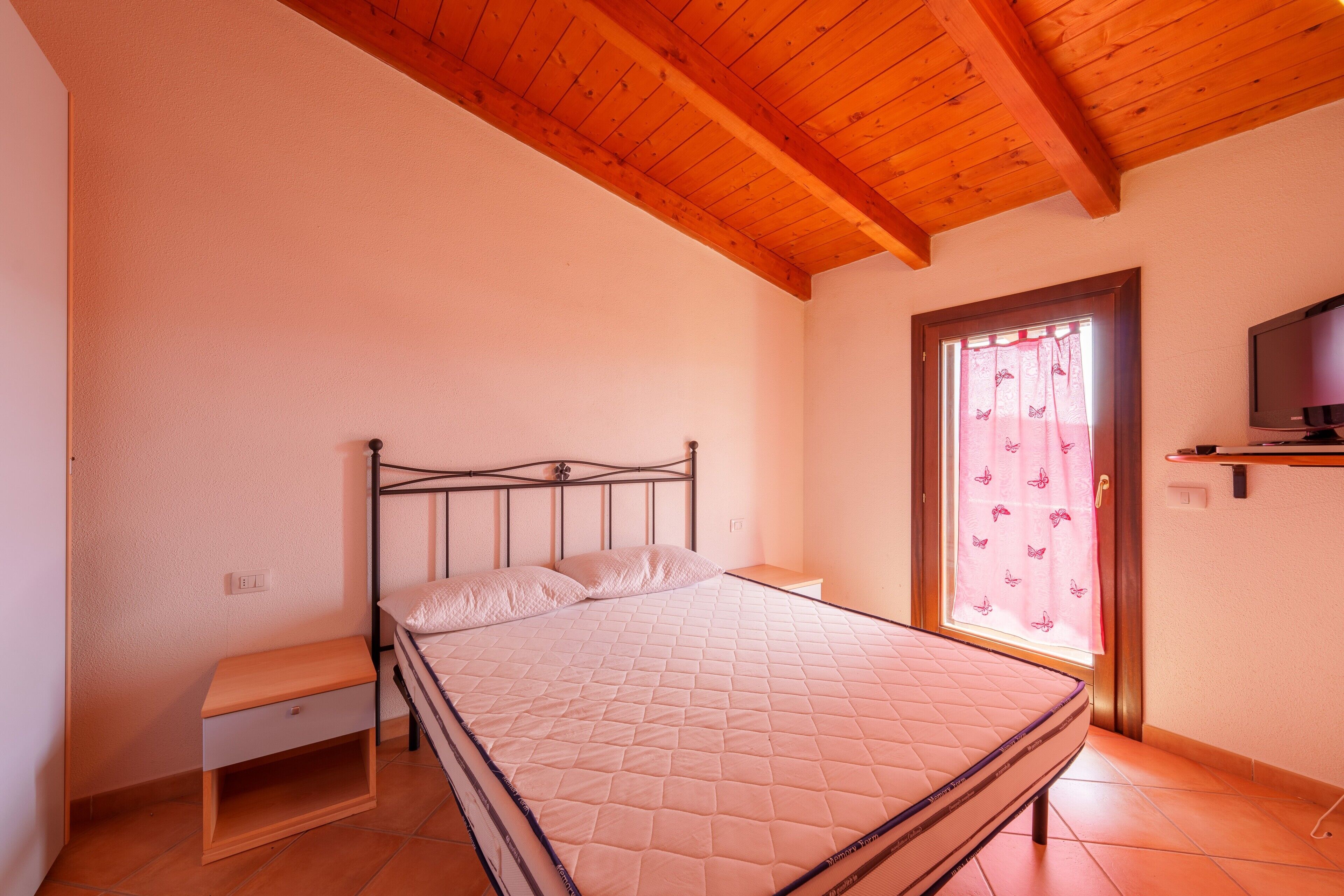 Une chambre à coucher avec un lit, une table de chevet, un téléviseur et une fenêtre avec des rideaux.