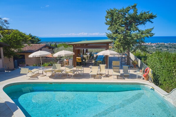 Una piscina con sillones de playa y sombrillas, un área de descanso cubierta y vista al mar.