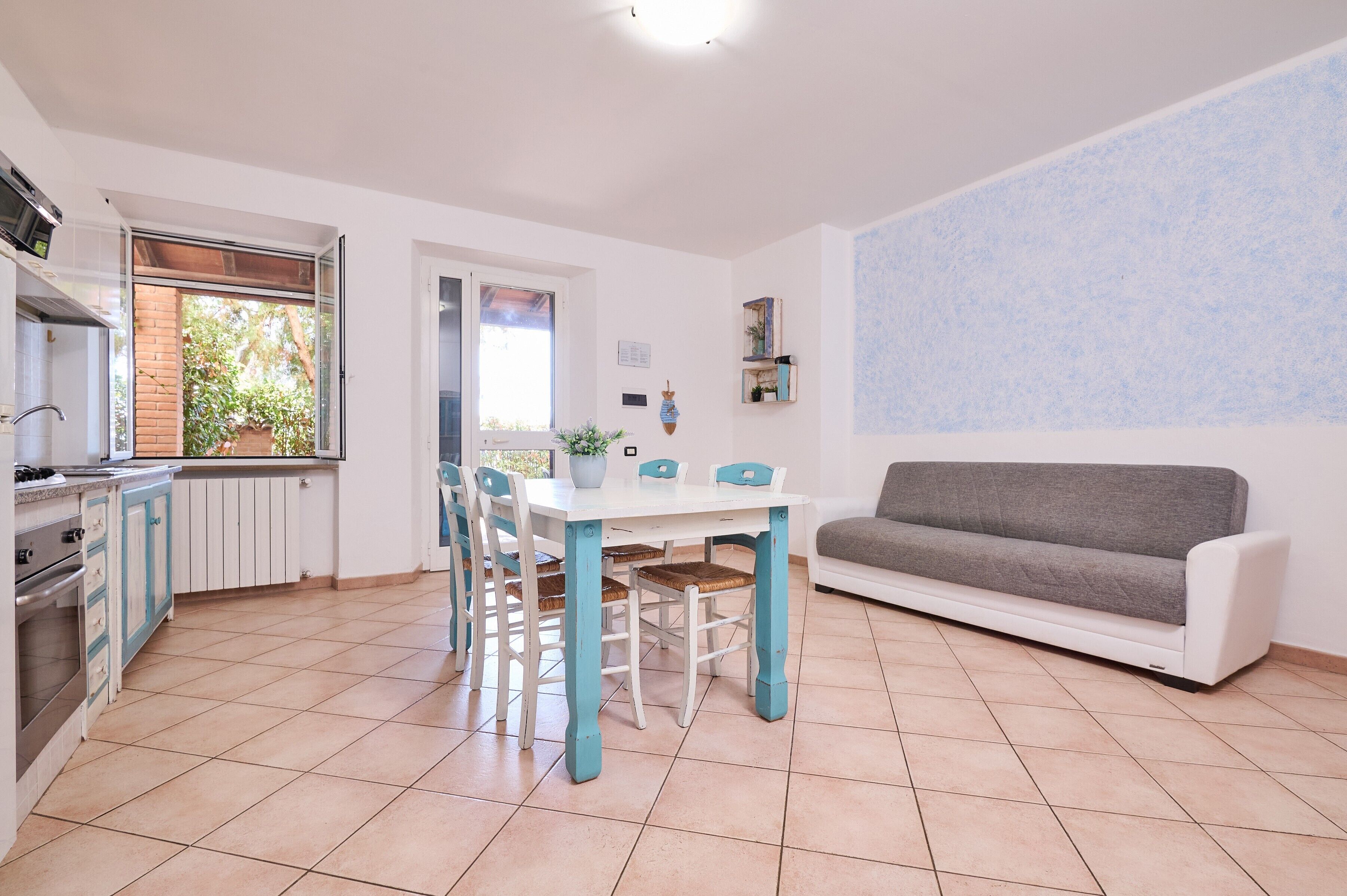 Un'area pranzo con tavolo bianco e sedie blu, un divano grigio e una cucina con finestra.