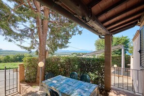 Un espace extérieur abrité, avec une table et des chaises, d’où l’on peut admirer un arbre et le paysage, et où l’on aperçoit un bâtiment au loin.