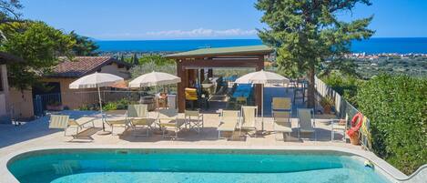 A swimming pool with lounge chairs and umbrellas, a covered seating area, and a view of the sea.