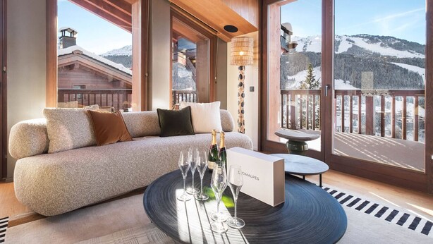 Ein modernes Wohnzimmer mit einer Couch, einem runden Glastisch mit Sektgläsern und Blick auf verschneite Berge durch große Fenster.
