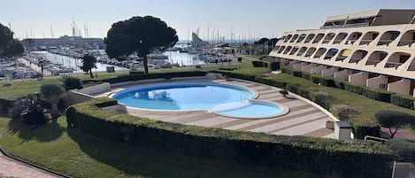 Una piscina con diseño circular, rodeada de setos cuidados y con vista despejada a un puerto deportivo al fondo.