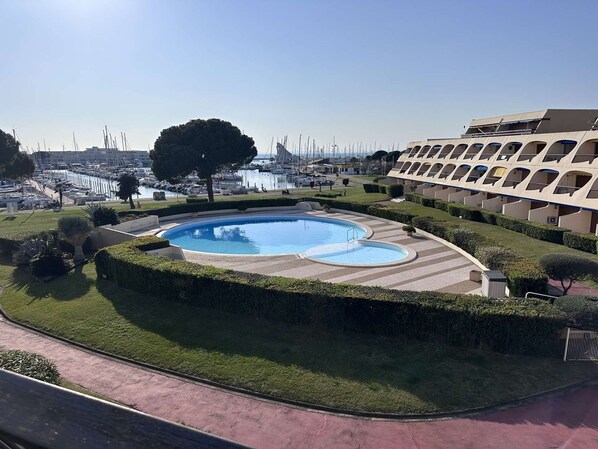 A swimming pool with a circular design, surrounded by well-maintained hedges and a clear view of a marina in the background.