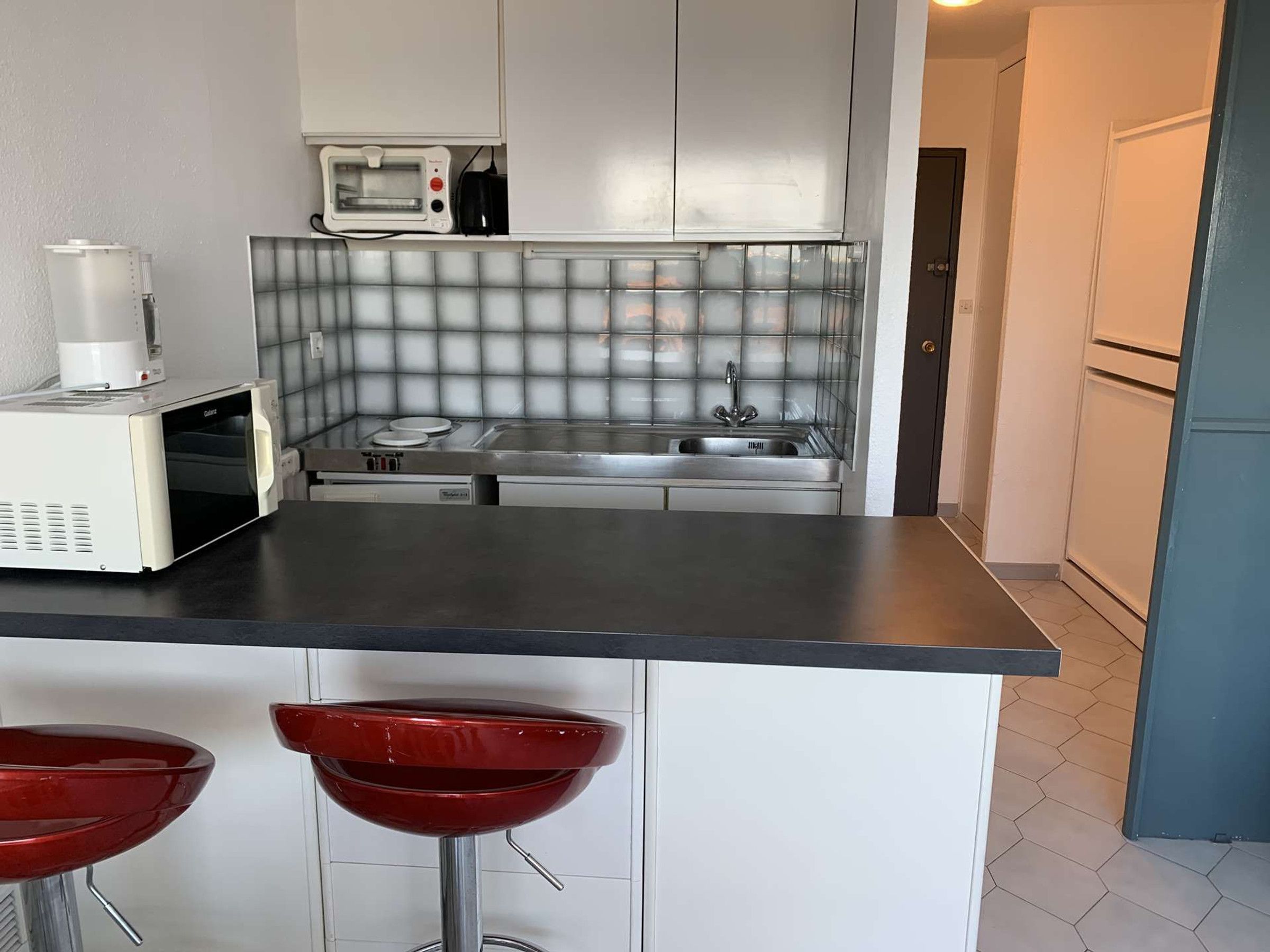 A compact kitchen with a microwave, stove, and sink. There are two red bar stools.