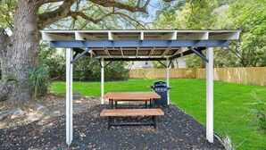 A covered outdoor seating area with a table and benches, surrounded by a wooden fence and a tree. - Beautiful Kitchen, BBQ, Outdoor Dining, Big Yard (St. Augustine)