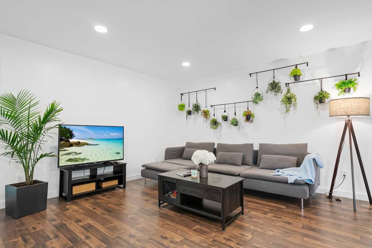 Een moderne woonkamer met een flatscreen televisie, een grijze bank, een salontafel en een plant in pot.