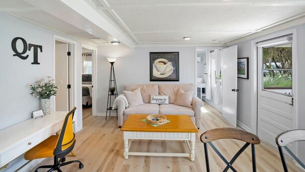 Ein modernes Wohnzimmer mit einem weißen Sofa, einem Couchtisch und einem Schreibtisch mit Stuhl. An der Wand hängt ein gerahmtes Bild und eine Tür mit Fenster.
