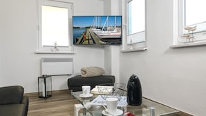 A modern living room with a glass coffee table, a sofa, a TV displaying a marina scene, and a window with a view of boats. - Bungalow LAETITIA - In a peaceful central location in Timmendorf Strand. (Insel Poel / OT Timmendorf Strand)