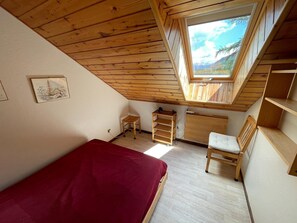 Ein Zimmer mit schrägen Wänden, einem Oberlicht, einem Bett, einem Sessel, einem kleinen Tisch und einer Kommode.