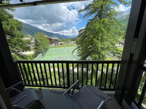 Ein Balkon mit Blick auf einen Tennisplatz, umgeben von Bäumen und Bergen.