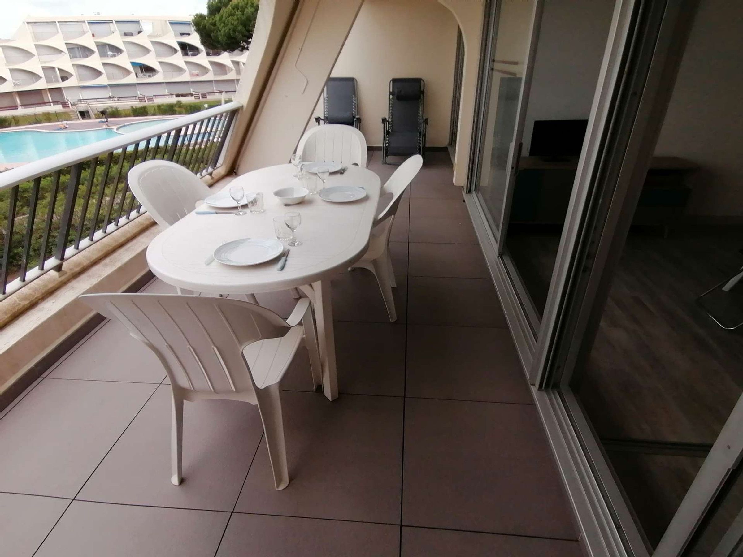 A balcony with a dining table set for two, chairs, a pool, and a building with a unique architectural design in the background.