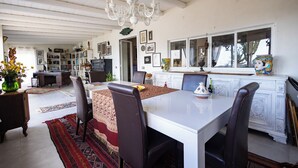 Un coin repas avec une table blanche, des chaises marron et un lustre. On aperçoit en arrière-plan une cuisine avec des placards et une fenêtre.