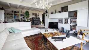 Un salon spacieux avec un canapé blanc, une table basse et un téléviseur. On y trouve également des étagères, un lustre et divers objets de décoration.