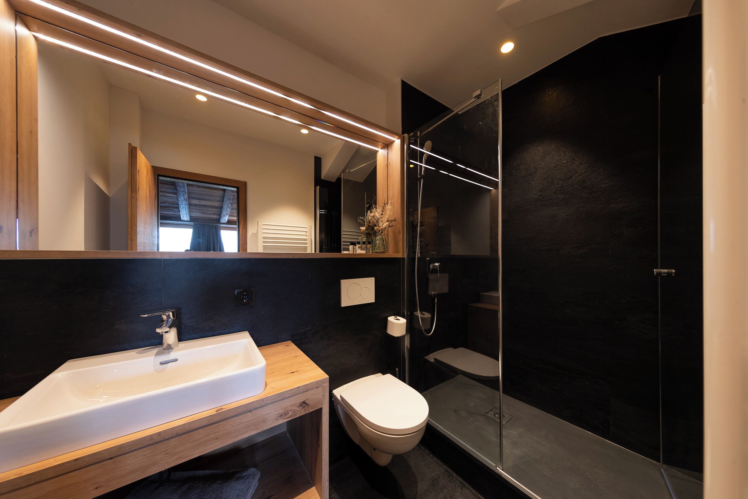 Un baño moderno con un mueble de tocador de madera, un lavabo blanco, un inodoro y una ducha con mampara de vidrio.