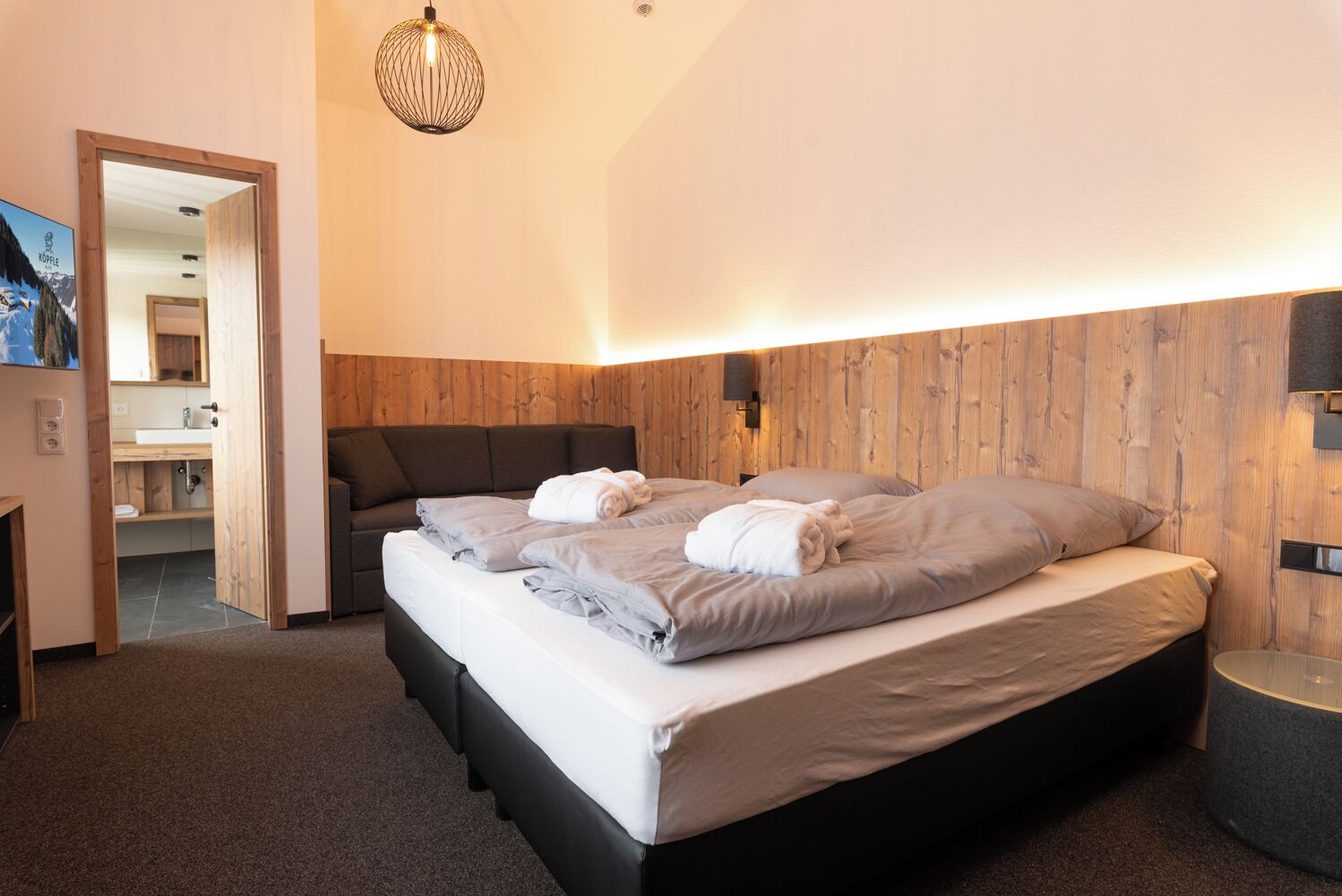 Une chambre d’hôtel moderne avec un mur d’accent en bois, un grand lit avec une literie grise, un petit canapé et une suspension.