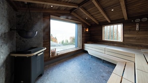 A rustic cabin interior with a stone wall, wooden beams, a large window, a wooden bench, and a trash bin. - Köpfle Alpe (Balderschwang)