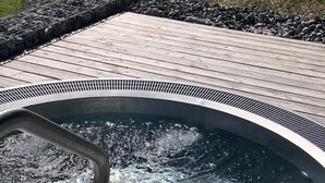A hot tub with a wooden deck and a stone border, surrounded by a grassy hillside and a wooden building in the background. - Köpfle Alpe (Balderschwang)