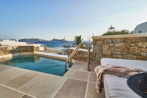 A pool area with a stone wall, a lounge chair, and a view of the sea and ships. - Aqua Mykonos Suites - Adults Only (Mykonos)