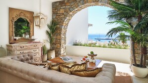 A cozy living room with a stone archway, a wooden dresser, a sofa, and a view of the sea. - Aqua Mykonos Suites - Adults Only (Mykonos)